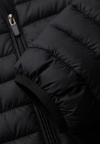 Detalle de una chaqueta acolchada negra, con textura de acolchado, puños elásticos y una cremallera. El material parece brillante y aislante.