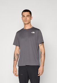 T-shirt grigia a girocollo realizzata in tessuto liscio. Presenta un piccolo logo bianco sul petto e maniche corte. Indossata con pantaloni scuri.