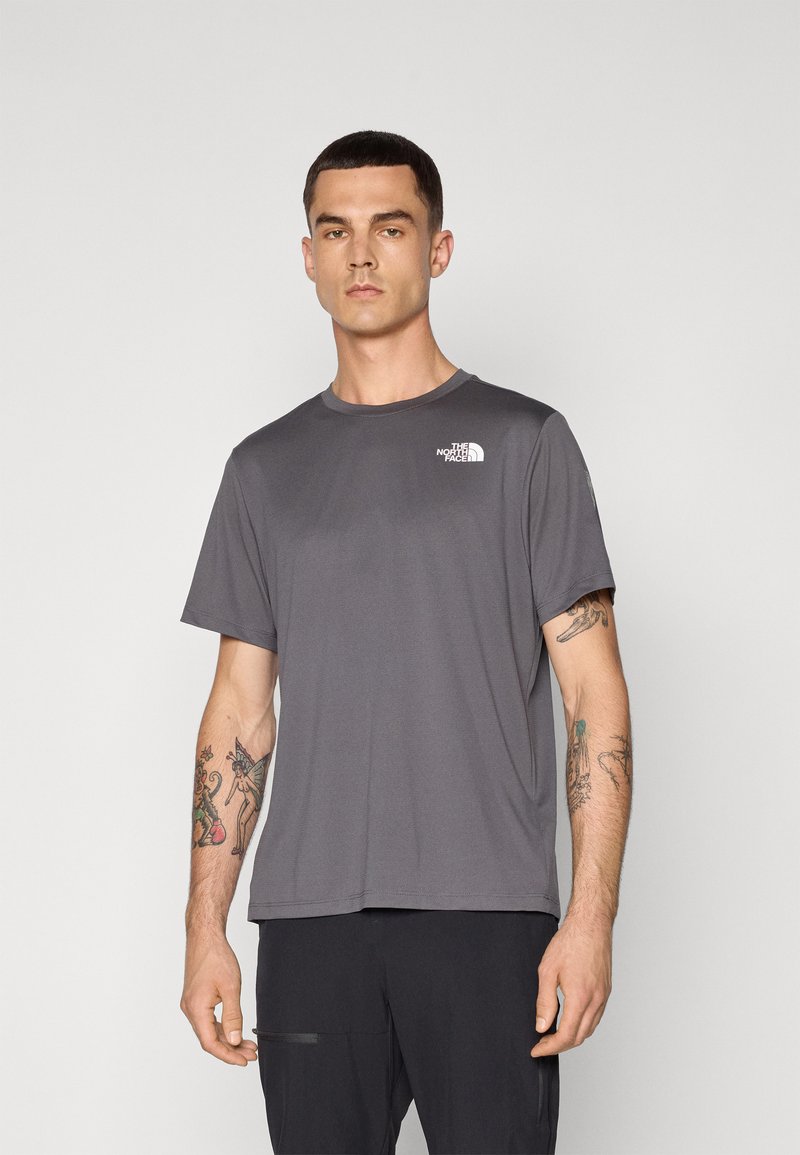 T-shirt grigia a girocollo realizzata in tessuto liscio. Presenta un piccolo logo bianco sul petto e maniche corte. Indossata con pantaloni scuri.