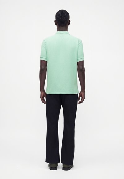 Homme vu de dos portant un polo vert clair à manches courtes, pantalon noir et chaussures foncées sur fond blanc uni.