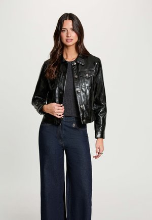 Donna con capelli lunghi e scuri che indossa una giacca di pelle nera sopra a un maglione scuro e jeans scuri a gamba larga, in piedi davanti a uno sfondo semplice.