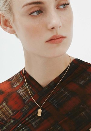 Collar de pendiente de oro con superficie texturizada, llevado sobre una cadena delgada, sobre una prenda con un diseño de patrón rojo y negro y escote en V.