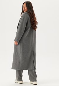 Cappotto lungo grigio con design dritto, spacchi laterali e tessuto testurizzato. Abbinato a pantaloni a gamba larga grigi e calzature chiare.