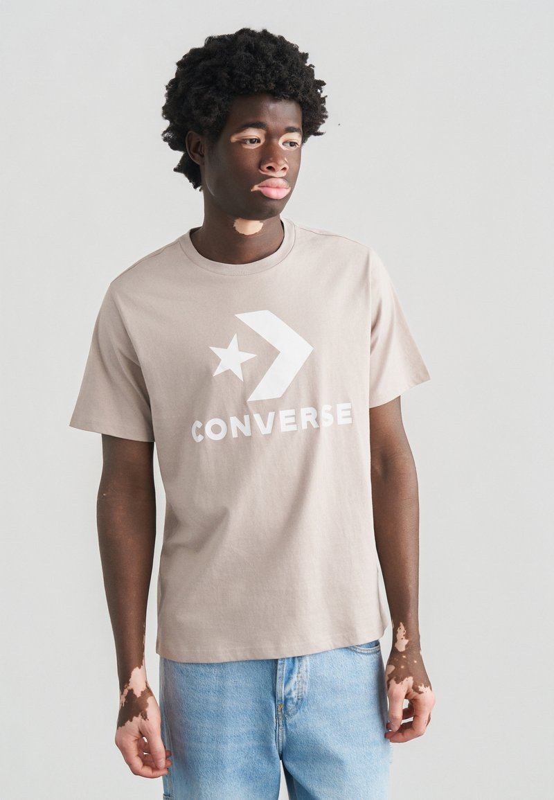 Μπεζ βαμβακερό μπλουζάκι με μεγάλο λευκό λογότυπο Converse που περιλαμβάνει σχέδιο αστέρι και βέλος. Συνδυάζεται με ανοιχτό μπλε τζιν.