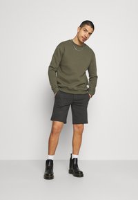 Olive green sweatshirt with ribbed cuffs, paired with dark gray shorts. Black ankle boots, white socks, and a silver chain necklace complete the look.