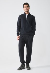 Hanorac negru cu fermoar, dotat cu o buzunar frontal, combinat cu pantaloni jogger asortați și pantofi sport negri, toate realizate dintr-un material moale și texturat.