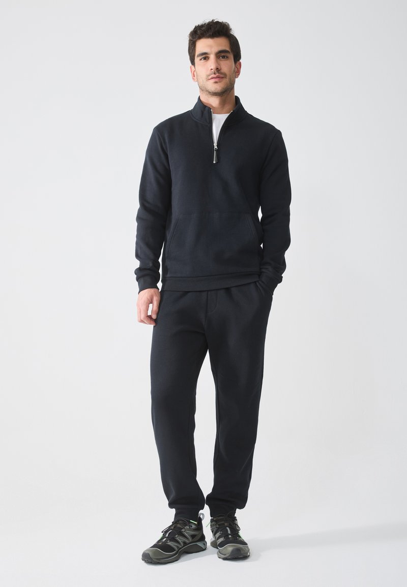 Hanorac negru cu fermoar, dotat cu o buzunar frontal, combinat cu pantaloni jogger asortați și pantofi sport negri, toate realizate dintr-un material moale și texturat.