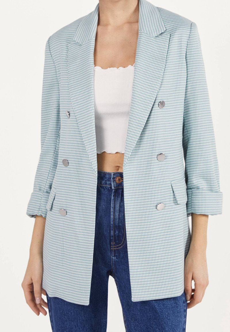 Femme portant un blazer croisé bleu clair à motif pied-de-poule, un top blanc court et un jean bleu taille haute, les mains reposant sur les côtés.