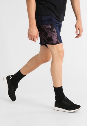 Ein Mann geht und trägt schwarze und lila gemusterte Sportshorts, schwarze Socken und schwarze Laufschuhe mit weißen Sohlen.