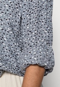 Chemise gris à motifs avec des éclaboussures noires et blanches, manches retroussées avec détail de bouton, fabriquée en tissu léger, mettant en avant une coupe décontractée.
