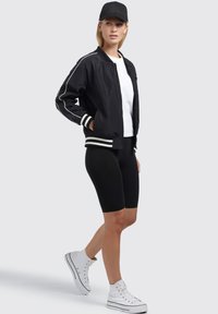Crna bomber jakna s bijelim trakama, bijela majica, crne biciklističke kratke hlače i bijeli visoki tenisice. Model nosi crnu kapu.