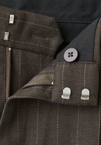 Prim-plan al unui articol vestimentar maro cu dungi fine, prevăzut cu fermoar, nasture și două închizători metalice tip cârlig și ochi pe o căptușeală de material negru.