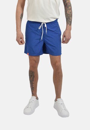 Uomo che indossa pantaloncini azzurri acceso con coulisse, sneakers bianchi e una maglietta bianca, in piedi con tatuaggi visibili su braccia e gamba sinistra.