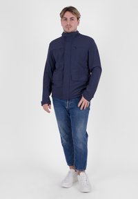 Giacca blu navy con colletto alto, chiusura con zip, due tasche sul petto e polsini elastici. Indossata con jeans azzurri chiari e sneakers bianche.