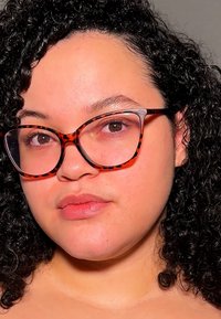 Occhiali tartarugati con forma oversize a occhio di gatto, montatura trasparente e dettagli sottili sulle asticelle. Capelli ricci di lunghezza spalla e trucco naturale.