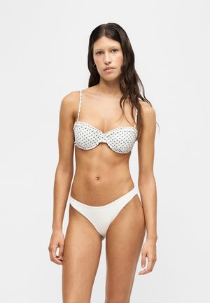 Femme aux longs cheveux foncés portant un bikini blanc avec un haut à pois noirs et un bas blanc uni, debout devant un fond uni.