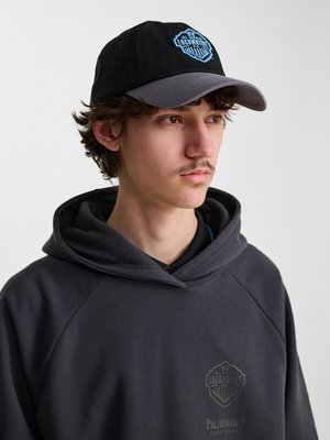 Młody mężczyzna z ciemnymi włosami i wąsami nosi czarno-szarą czapkę baseballową i czarną bluzę z kapturem z logo "El Paso Locomotive FC", zwrócony w prawą stronę.