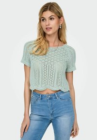 Lys mintgrøn strikket crop top med korte ærmer, der har intrikate blonder og en bølget kant, sammen med blå højlivede jeans.