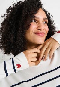 Mujer sonriente con cabello rizado, vestida con un suéter blanco con rayas marinas y detalles de corazones rojos en las mangas, posando con las manos cerca de su cara.