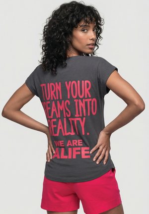 Frau mit lockigem dunklem Haar blickt über ihre Schulter, trägt ein graues Shirt mit rotem Text "TURN YOUR DREAMS INTO REALITY. WE ARE ALIFE" und pinke Shorts.