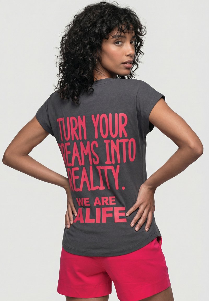 Frau mit lockigem dunklem Haar blickt über ihre Schulter, trägt ein graues Shirt mit rotem Text "TURN YOUR DREAMS INTO REALITY. WE ARE ALIFE" und pinke Shorts.
