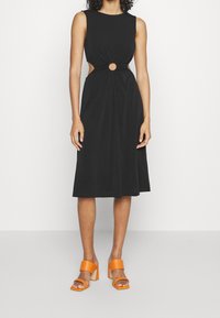 Robe noire sans manches avec un design découpé mettant en avant un accent circulaire à la taille ; assortie avec des sandales à talons carrés orange.