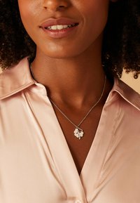 Collier en chaîne en argent avec un pendentif en trèfle à quatre feuilles, porté avec une chemise en satin rose clair, mettant en avant une surface polie et lisse.