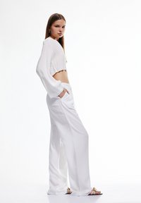 Witte cropped langmouwen top met gegolfde zoom, gecombineerd met losvallende witte broek. Model poseert in sandalen, waarbij een minimalistisch ontwerp wordt getoond.