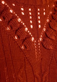 Gros plan sur un tissu tricoté rouge avec des motifs complexes de câbles et de dentelle formant des dessins géométriques texturés.