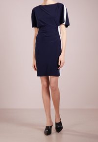 Kvinna klädd i en marinblå knälång klänning med vita sidostycken och svarta spetsiga mules, stående på ett ljusbeige golv.