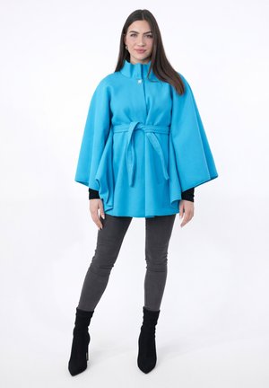 Femme portant une cape manteau bleu vif ceinturée, un haut noir, un jean skinny gris et des bottines à talons noires, debout sur un fond blanc.