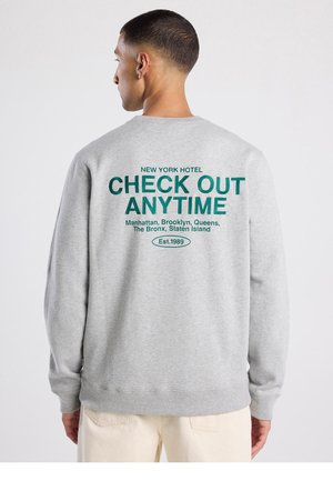 Grey sweatshirt featuring bold green text on the back: "NEW YORK HOTEL CHECK OUT ANYTIME" with city names and "Est. 1989."