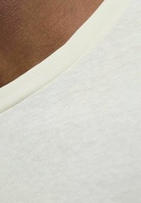 Balts adīts audums ar gludu tekstūru, tuvplānā redzama apaļa kakla izgriezuma daļa un tumša ādas krāsa fonā.