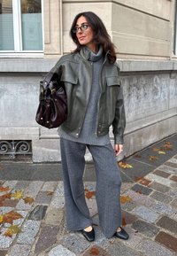 Nainen harmaa neulepaita päällään ja oliivinvärinen nahkatakki yllään pitelee tummaa ruskeaa käsilaukkua, seisoo mukulakivikadulla, jolla on pudonneita lehtiä.