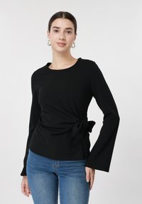 Νεαρή γυναίκα με σκούρα μαλλιά φοράει μαύρο μακρυμάνικο τοπ δεμένο στη μέση και μπλε τζιν, ποζάροντας μπροστά σε λευκό απλό φόντο.