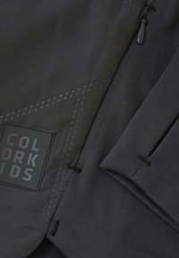 Svart tygjacka med en slät textur, en upphöjd logotyp-patch och subtila prickiga mönster. Inkluderar stickningsdetaljer och en dragkedja.