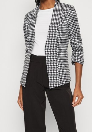 Vrouw draagt een zwart-wit pied-de-poule blazer over een wit topje met zwarte hoge taillebroeken, cropped bij de schouders en taille.