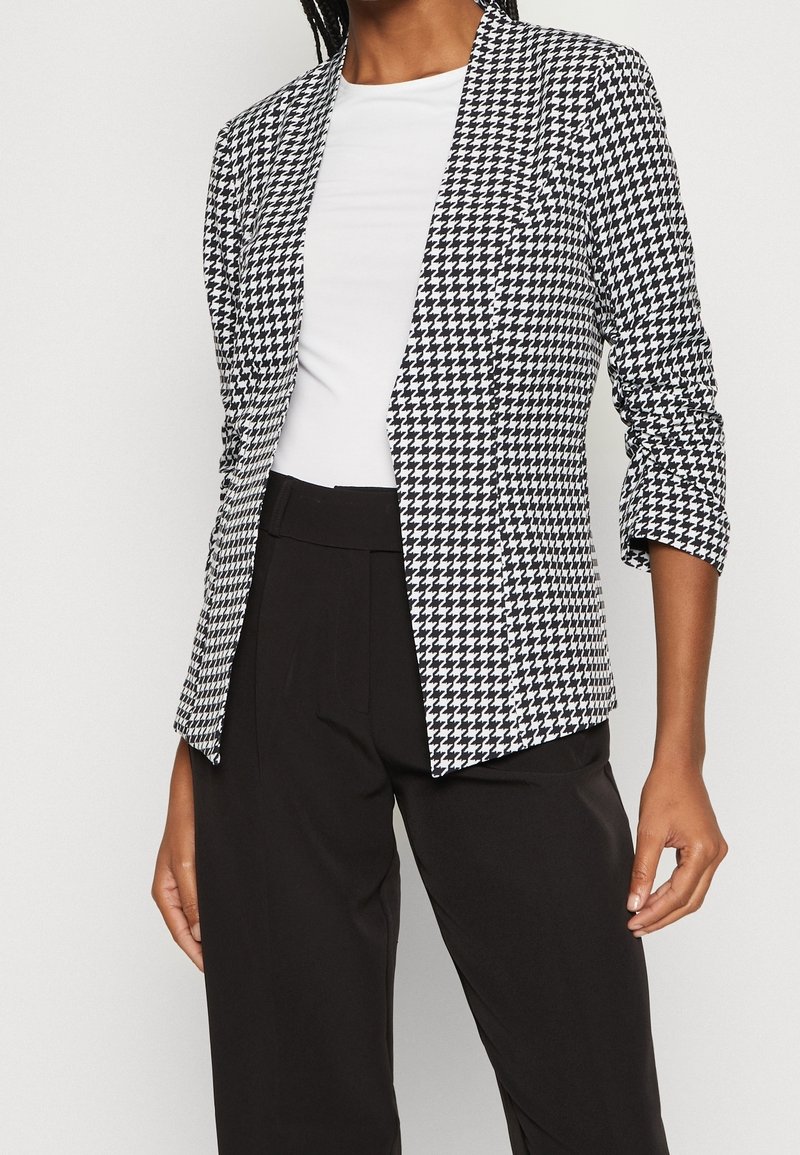 Femme portant un blazer pied-de-poule noir et blanc par-dessus un top blanc avec un pantalon noir taille haute, raccourci aux épaules et à la taille.