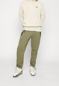 Beige prickig hoodie med dragsko, lager på en ljusgrön skjorta. Oljefärgade tapered joggers med svarta dragkedjefickor. Vita sneakers.
