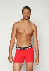 Rote Boxershorts mit schwarzem elastischem Bund, auf dem das "ellesse"-Logo zu sehen ist. Der Stoff wirkt glatt und dehnbar. Das Modell ist oberkörperfrei und steht aufrecht.