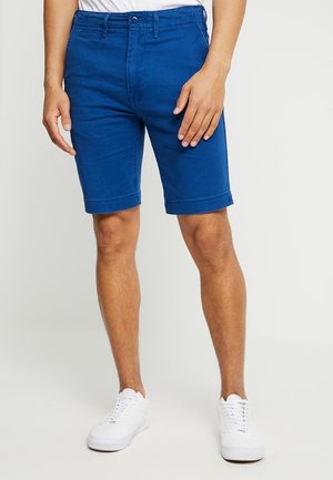 Homme portant un short bleu au niveau des genoux et des baskets blanches, debout devant un fond clair uni.