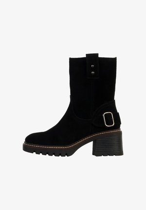 Bottine en suede noir avec un talon épais, accent en boucle et une semelle cousue. Dispose d'une pointe plate et d'une sangle décorative au niveau de la cheville.
