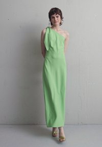 Φόρεμα μέχρι το πάτωμα με έναν ώμο σε ζωηρό πράσινο ύφασμα, που διαθέτει φαρδύ φιόγκο στον ώμο. Συνδυάζεται με λεπτά, ανοιχτόπατα τακούνια.