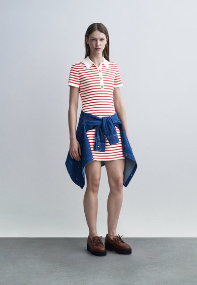 Γυναίκα που φοράει κόκκινο και λευκό φόρεμα με ρίγες και κοντά μανίκια, με λευκό γιακά, μπλε μπουφάν δεμένο στη μέση και καφέ πλατφόρμες παπούτσια.