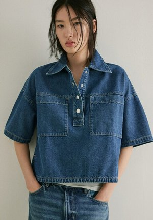 Jeune personne aux longs cheveux foncés portant une chemise en denim bleu ample à manches courtes et grandes poches avant, les mains dans les poches du jean bleu.
