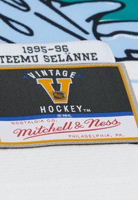 Vintage NHL Eishockey-Trikot-Patch mit schwarzem Hintergrund, gelbem "V" und roter "Mitchell & Ness" Schrift. Weißer Stoffuntergrund mit Etiketten.