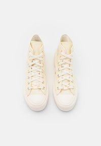 Vysoké tenisky v off-white plátně s bílými tkaničkami, gumovou špičkou, mosaznými očky a hladkým texturem. Klasický design s minimálním zdobením.