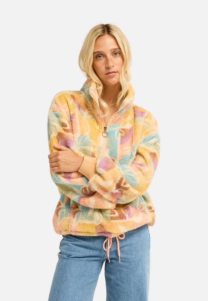 Donna con capelli biondi lunghi indossa una giacca in pile colorata con motivo pastello e mezza cerniera, e jeans azzurri chiari su sfondo semplice.
