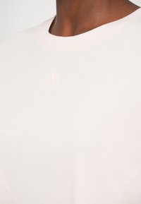 Tricou din bumbac roz deschis, cu guler rotund și un logo subtil embosat, care prezintă "AX" pe piept. Textură fină, design minimalist.