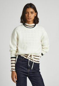 Maglione bianco a trecce con polsini a coste sopra un dolcevita a righe, abbinato a jeans scuri con vita regolabile e dettagli con logo.